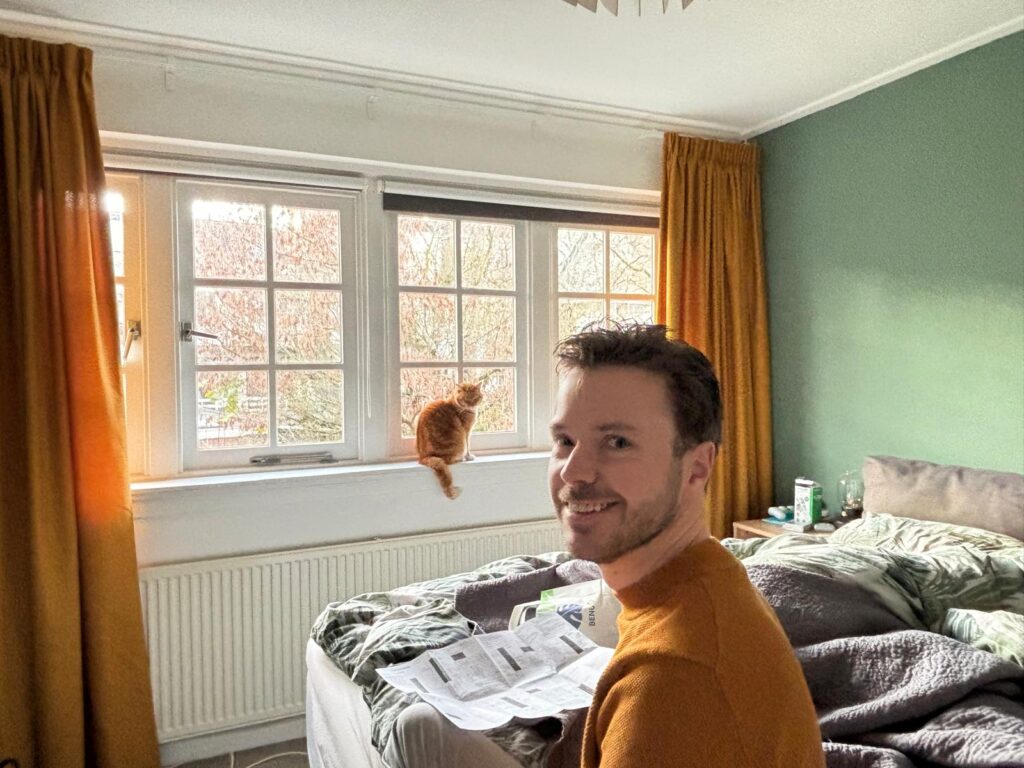 Stefan, de schrijver van deze blog, kijkt naar de camera. Hij leunt op het bed. Achter hem op de vensterbank zit zijn kat Charlie.